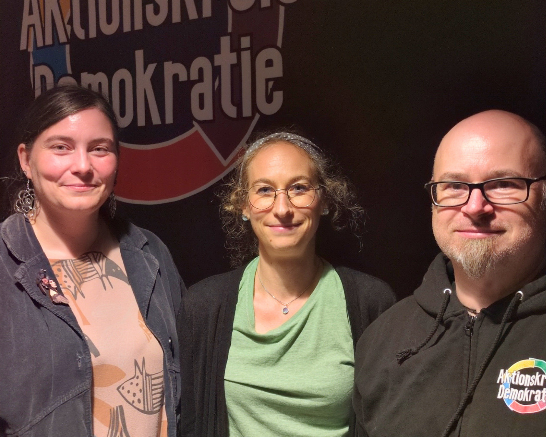 Vor einem gro&szlig;en Logo des AKtionskreis Demokratie stehen zwei Frauen und ein Mann. Es handelt sich um die neuen Vereinsvorsitzenden Alexandra Seefeld, Anna Michaelis und Knut Meenzen